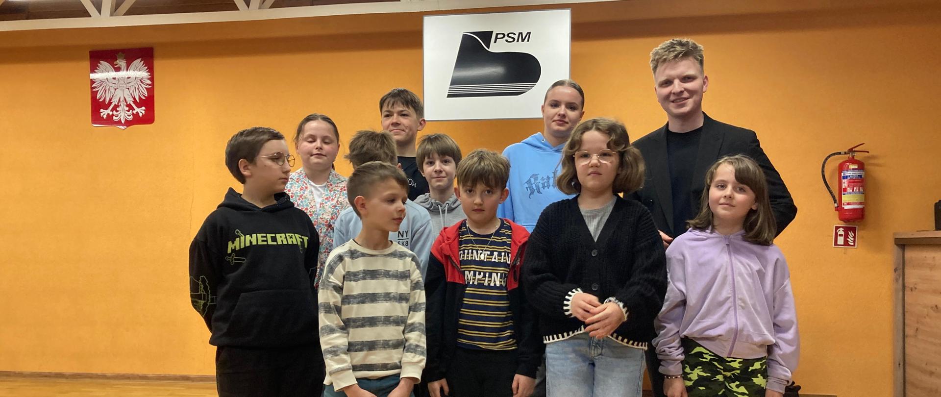 16 kwietnia 2024 r - odbyły się warsztaty gitarowe z Mateuszem Kowalskim z Uniwersytetu Muzycznego Fryderyka Chopina w Warszawie. W auli naszej szkoły po koncercie uczniowie klasy gitary wraz z wykładowcą Mateuszem Kowalskim stoją na scenie. Wszystkich jest 11 osób - pan Mateusz stoi po prawej stronie