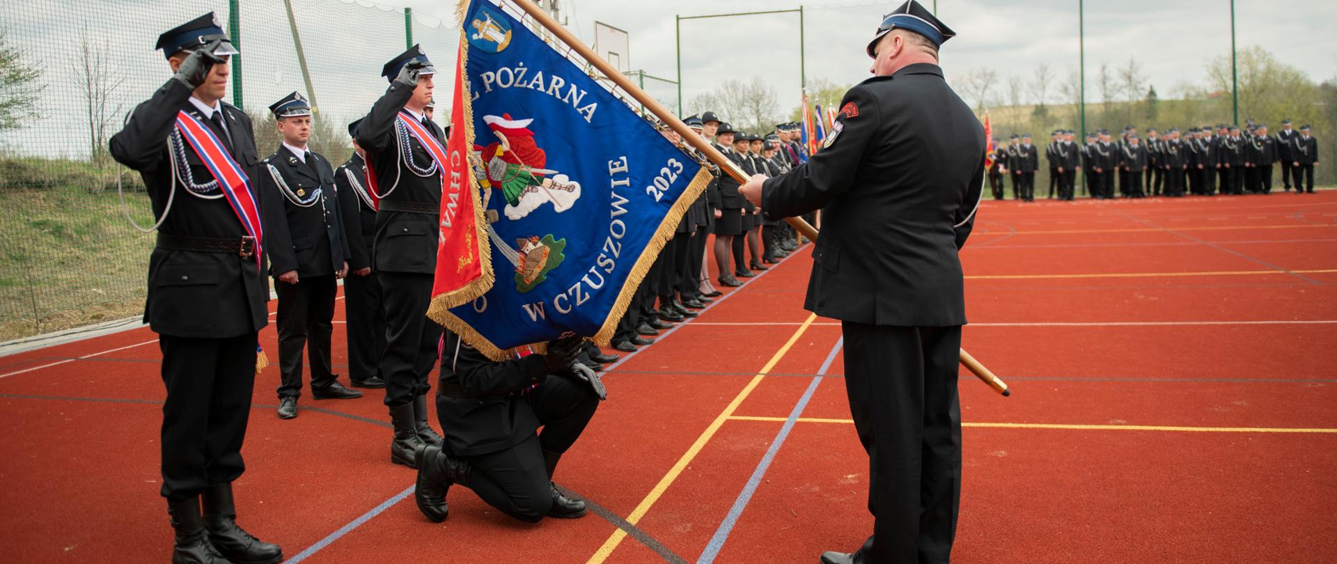 Na pierwszym planie mężczyzna trzyma sztandar na drugim planie mężczyzna klęcząc na jedno kolanu całuje płat sztandaru 