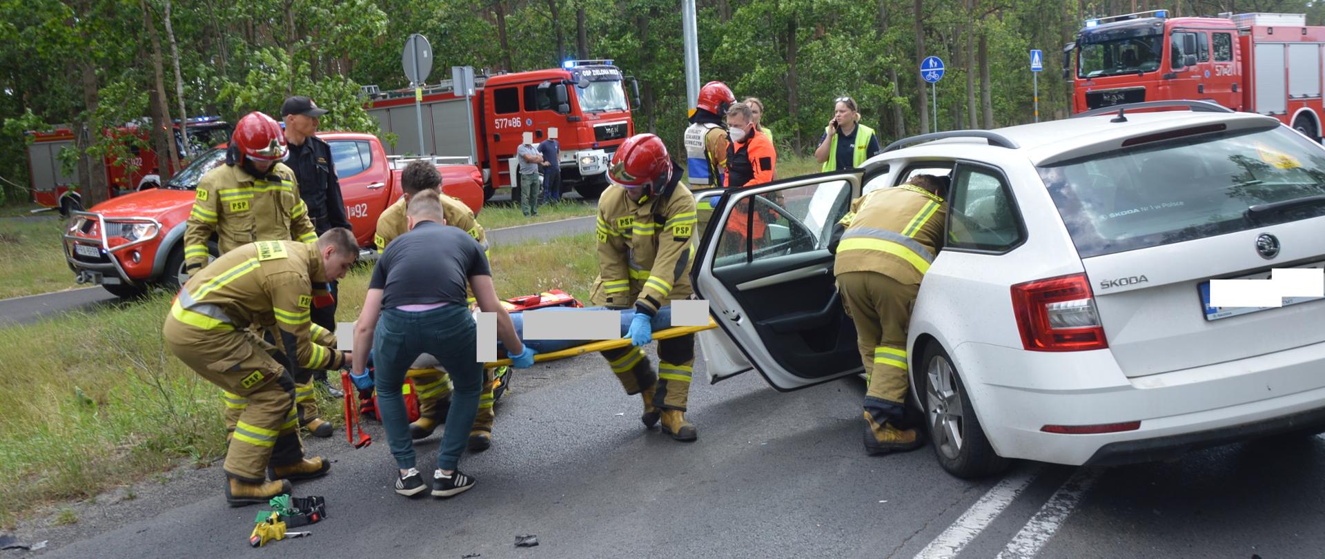 Zderzenie aut na skrzyżowaniu. Na pierwszym planie biała skoda octavia, z której strażacy ewakuują na desce poszkodowana kierującą. Koło pojazdu znajdują się też dwie policjantki i ratownik medyczny. Na tle lasu stoją cztery pojazdy straży pożarnej.