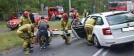 Zderzenie aut na skrzyżowaniu. Na pierwszym planie biała skoda octavia, z której strażacy ewakuują na desce poszkodowana kierującą. Koło pojazdu znajdują się też dwie policjantki i ratownik medyczny. Na tle lasu stoją cztery pojazdy straży pożarnej.