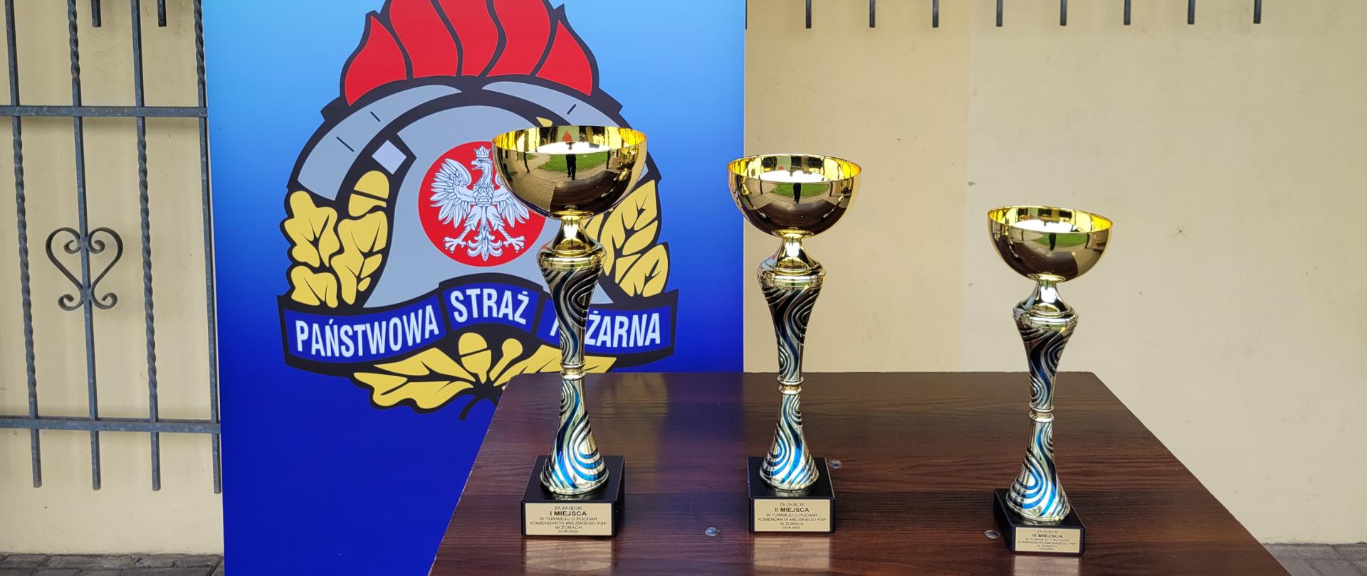 Puchary ustawione na stoliku na tle ścianki z logo PSP oraz napisem Komenda Miejska Państwowej Straży Pożarnej w Żorach.