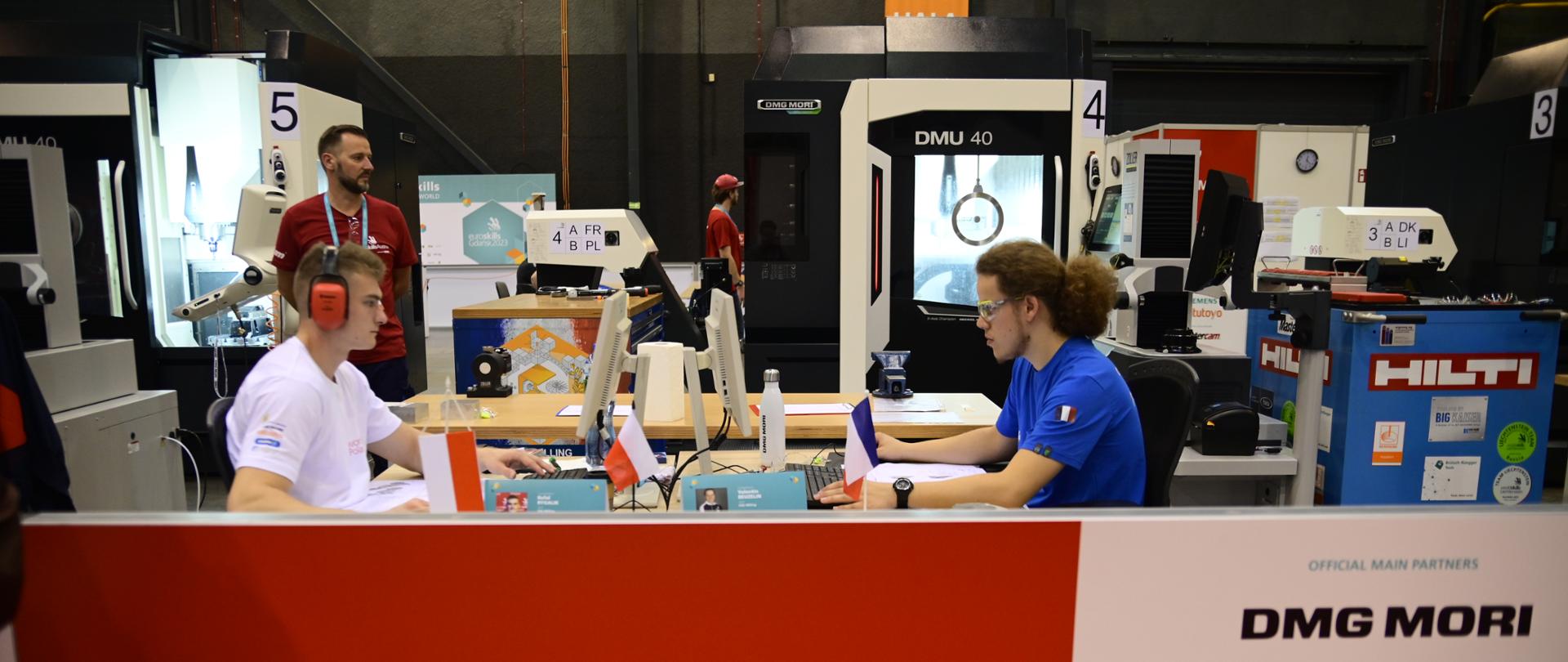 Widok na stanowisko, przy stoliku siedzi naprzeciwko siebie dwóch mężczyzn, przed nimi na czerwonej ściance napis CNC Milling.