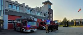 Przed budynkiem Państwowej Straży Pożarnej stoją samochody ratowniczo - gaśnicze i specjalny drabina z włączonymi sygnałami świetlnymi, przed samochodami w szeregu stoją w ubraniach koszarowych funkcjonariusze oddając hołd zmarłemu druhowi. Po prawej maszt z podniesioną flagą państwową