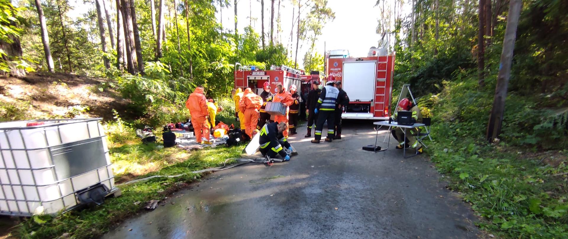 Zdjęcie przestawia pracę specjalistycznej grupy ratownictwa chemiczno-ekologicznego ze Skarżyska-Kamiennej. Strażacy przygotowują się do uszczelniania zbiornika i likwidacji wycieku substancji niebezpiecznej. Obok nich stoi zbiornik przygotowany do przepompowania substancji. Dookoła widać las mieszany.