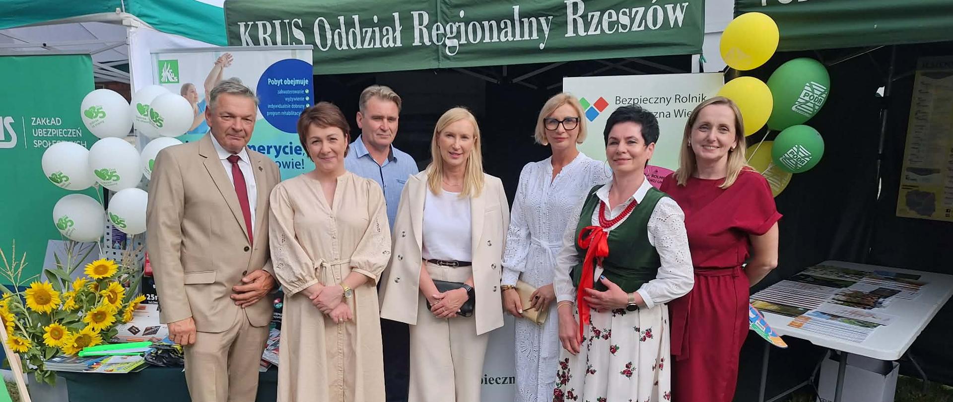Pięć kobiet i dwóch mężczyzn pozuje do zdjęcia przed stoiskiem Kasy pod zielonym namiotem KRUS. Z tyłu za nimi znajduje się stół, na którym są wyłożone materiały prewencyjne Kasy (odblaski, ulotki). Po lewej stronie stoją żywe słoneczniki.