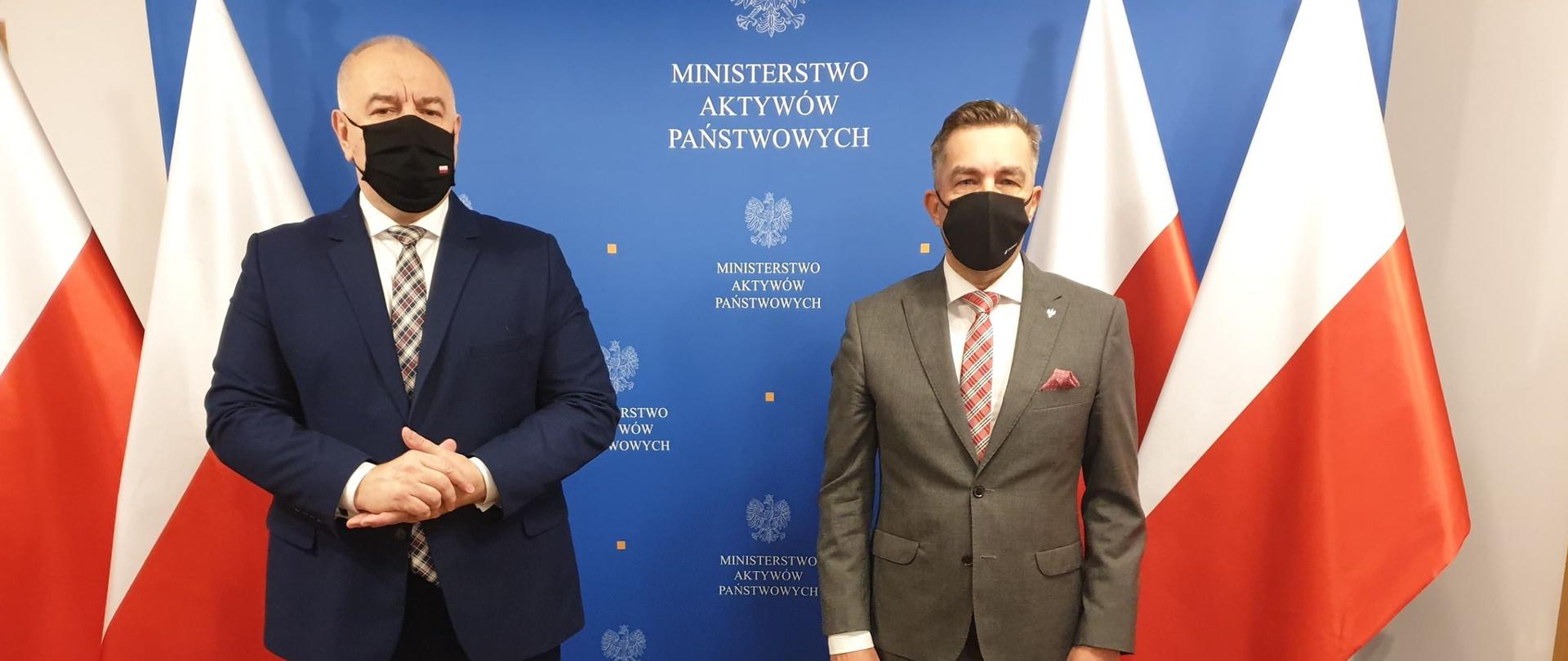 Wicepremier Jacek Sasin oraz wiceminister Zbigniew Gryglas na tle ścianki. Po obu stronach flagi Polski. 