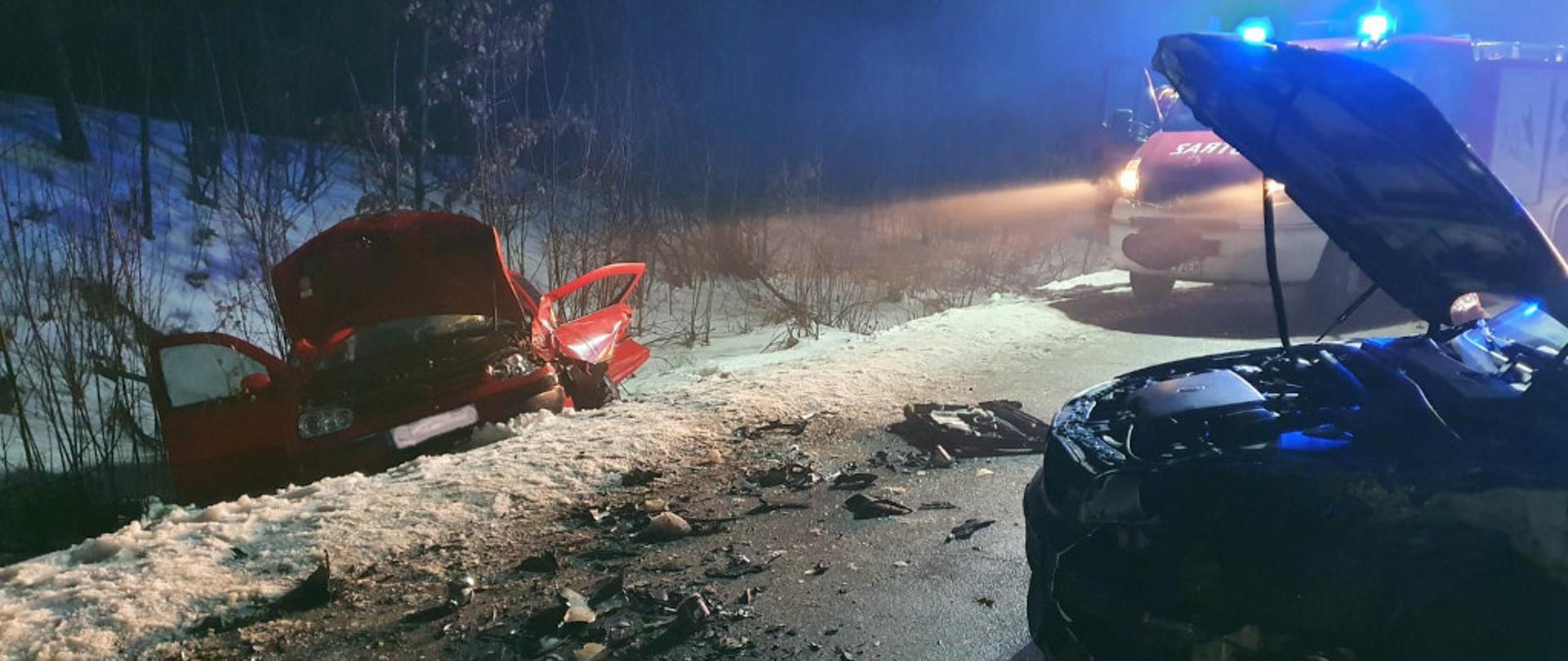 Zdjęcie przedstawia dwa rozbite samochody osobowe. Jeden z nich w kolorze czerwonym znajduje się w rowie z otwartą maską. Przód drugiego widniejący z prawej strony zdjęcia stoi na drodze również z otwartą maską oraz licznymi uszkodzeniami karoserii. W tle znajduje się pojazd pożarniczy z włączonymi błyskowymi światłami alarmowymi w kolorze niebieskim. Zdjęcie zrobione w porze nocnej. Na poboczu zalega biała warstwa śniegu.