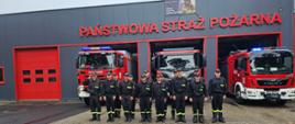 Na zdjęciu widoczny budynek komendy państwowej straży pożarnej, nad garażami napis: Państwowa Straż Pożarna. Przed budynkiem stoją w szeregu strażacy w czarnych ubraniach koszarowych. Za nimi ustawione w bramach garażowych trzy czerowne samochody specjalne.