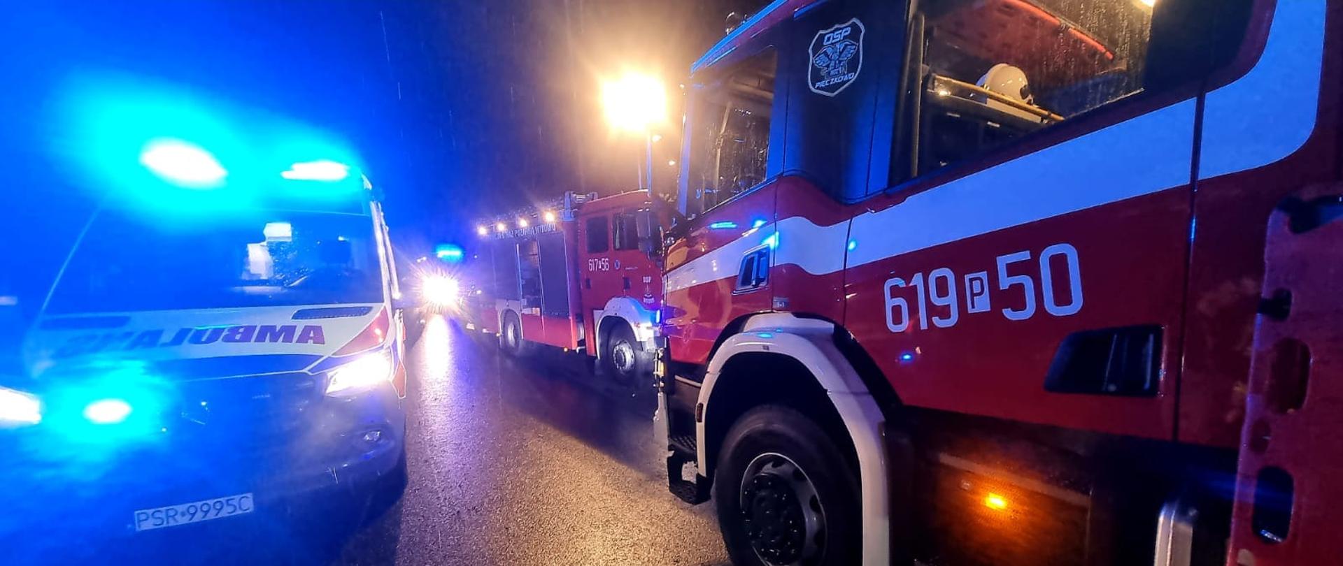 Samochody pożarnicze i zespół ratownictwa medycznego w czasie nocnej akcji poszukiwawczej. Samochody stoją na ulicy.