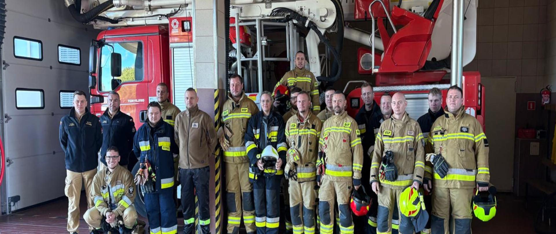 Grupa strażaków stoi na w pomieszczeniu remizy ubrani w ubrania specjalne koloru piaskowego i granatowego, trzymają w ręku hełmy. Strażacy stoją na tle samochodu specjalnego, podnośnika hydraulicznego. 