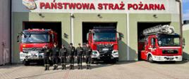 Zdjęcie przedstawia strażaków na tle samochodów oraz garaży jednostki ratowniczo-gaśniczej.