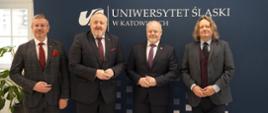 Na tle szarej ścianki z napisem Uniwersytet Śląski w Katowicach stoi czterech mężczyzn w garniturach, wiceminister Gzik trzeci od lewej, w tle z lewej strony okno.