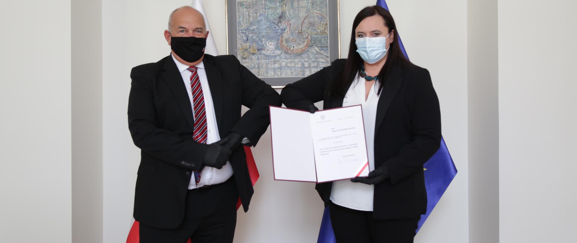 W pomieszczeniu na tle dwóch flag - polskiej i unijnej - stoi od lewej minister Tadeusz Kościński. Obok niego wiceminister Małgorzata Jarosińska-Jedynak, która prezentuje dokumenty z powołania jej na stanowisko sekretarza stanu.