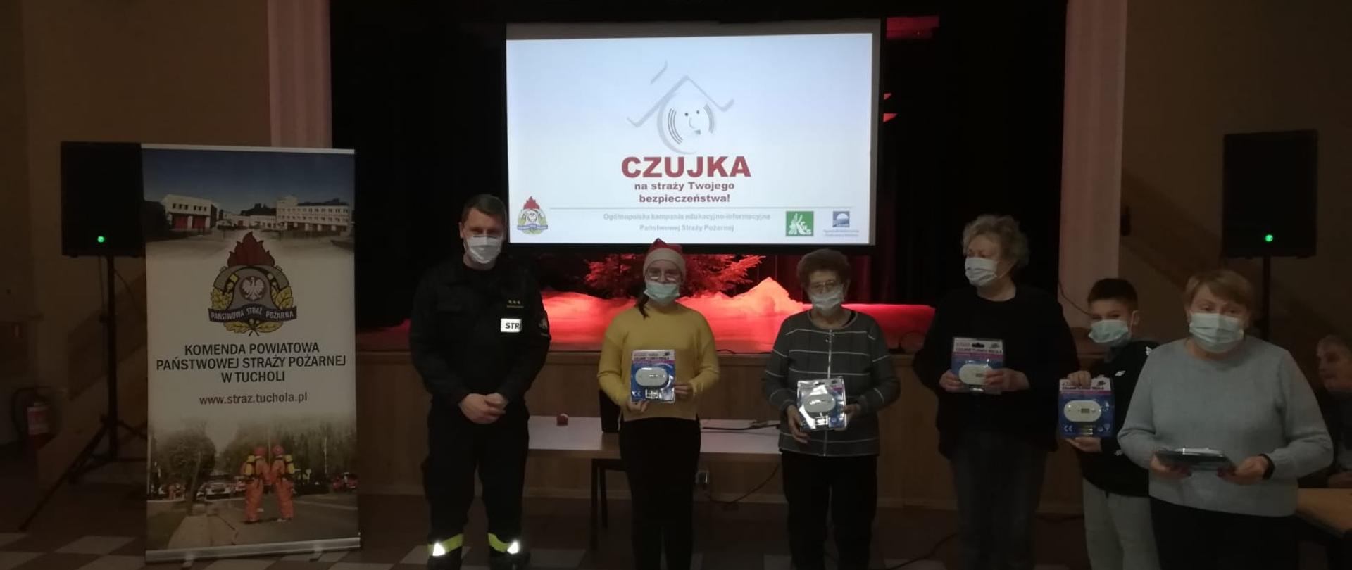 Zdjęcie przedstawia grupę osób, które wylosowały nagrody (czujki tlenku węgla) podczas prelekcji. Nagrodzeni stoją obok strażaka prowadzącego pogadankę. Na zdjęciu jest 5 osób wyróżnionych i strażak. W tle widać slajd prezentacji o czujkach tlenku węgla.