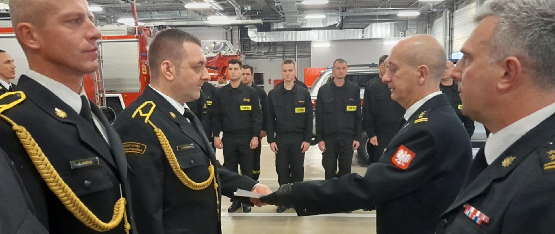 Komendant Miejski PSP wraz z Zastępcą wręczają awanse wyróżnionym strażakom. W tle strażacy oraz samochody ratowniczo - gaśnicze. 