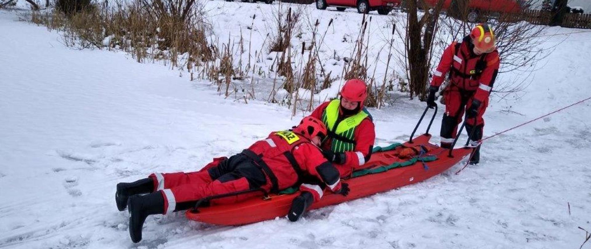 Na zdjęciu widać osobę osobę ratowaną, oraz ratowników przy brzegu jeziora. Ratownicy ubrani w kombinezony niezatapialne, poszkodowany znajduje się na desce lodowej. Obydwaj przypięci są linką asekuracyjną. 
