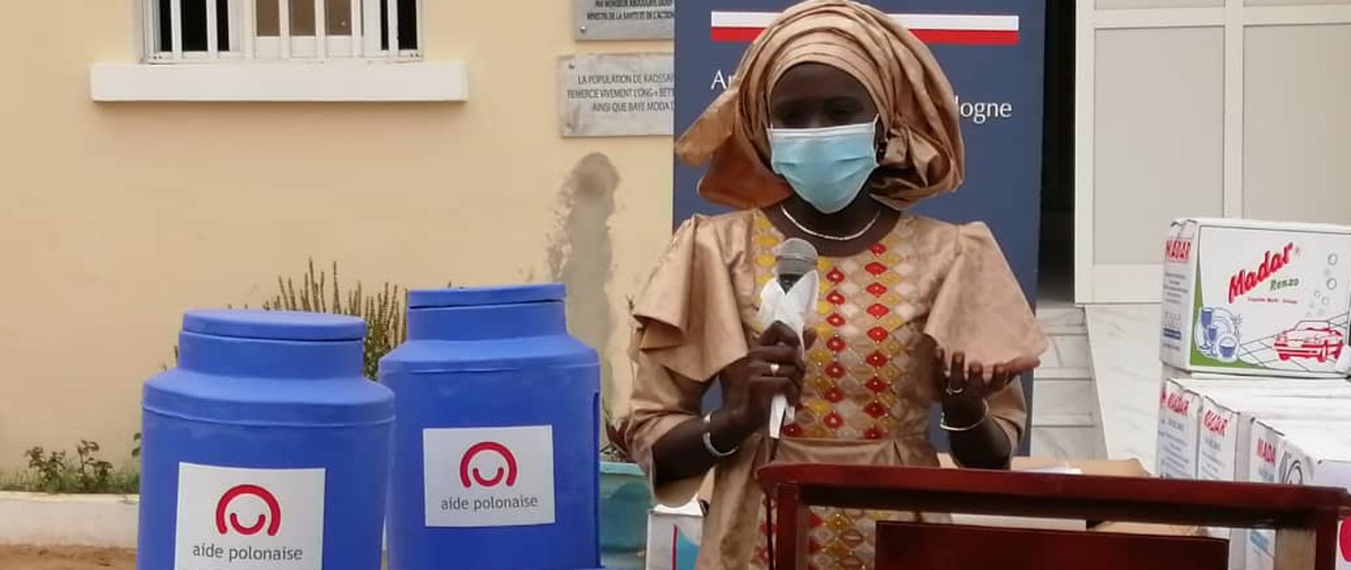 kobieta w tradycyjnym stroju trzyma mikrofon, przy niej stoją niebieskie pojemniki na wodę