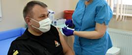 Na zdjęciu widoczny nadbrygadier Andrzej Babiec w umundurowaniu dowódczo-sztabowym poddający się szczepieniu. Personel szpitala w niebieskim fartuchu dokonuje wkłucia podając szczepionkę w lewe ramię siedzącego funkcjonariusza.Osoby na zdjęciu są w maseczkach oraz rękawiczkach.