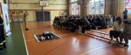 Zdjęcie przedstawia szkolenie z pierwszej pomocy prowadzone przez strażaka w sali gimnastycznej. Na pierwszym planie, na środku boiska, leży manekin do resuscytacji na macie, a obok niego stoi strażak w mundurze, który prawdopodobnie coś objaśnia. Liczna grupa młodzieży siedzi na ławkach i krzesłach, obserwując pokaz. W tle widoczne są okna, boisko do koszykówki oraz tablica wyników. Na lewo od sceny znajduje się baner Komendy Powiatowej Państwowej Straży Pożarnej w Zwoleniu. Sala jest udekorowana kolorowymi balonami.