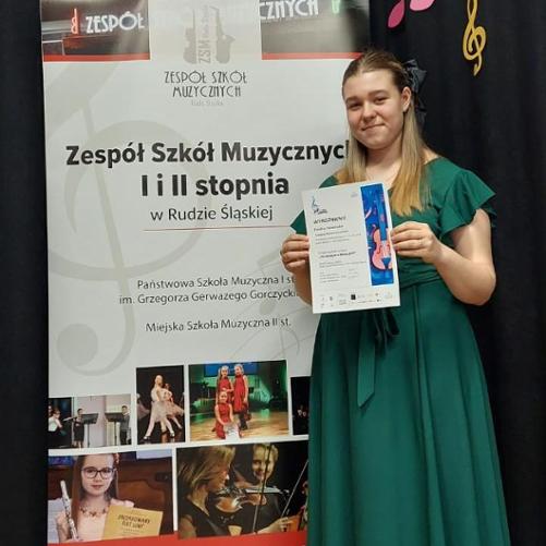 Nastolatka stoi przed banerem Zespołu Szkół Muzycznych I i II st. w Rudzie Śląskiej i trzyma w rękach dyplom.