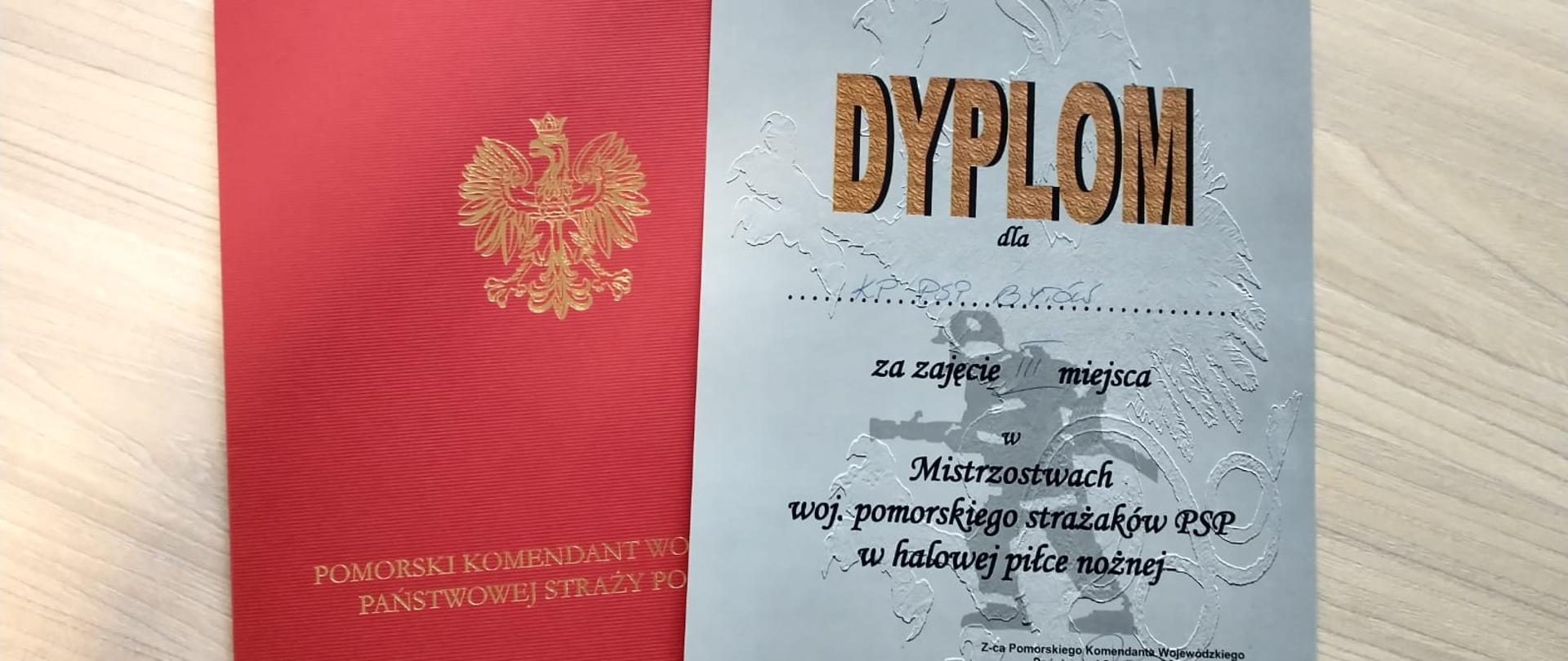 Dyplom za zajęcie III miejsca wraz z czerwoną teczką leżą na blacie biurka