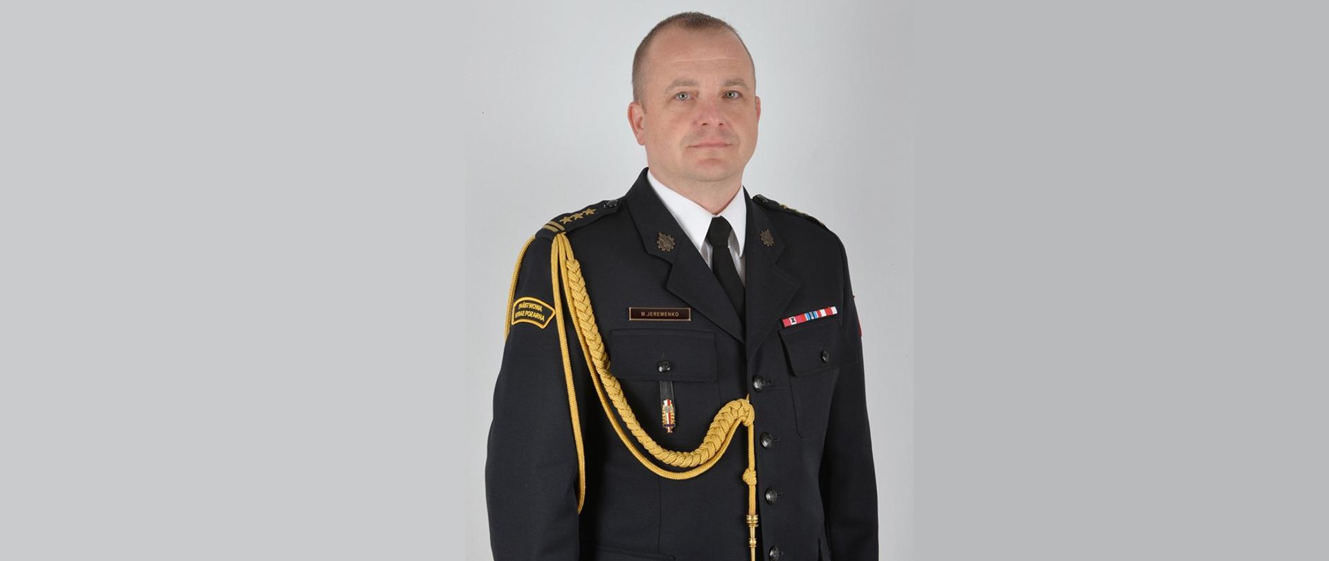 Portret funkcjonariusza Państwowej Straży Pożarnej w mundurze galowym. Mężczyzna stoi na jasnym tle, zwrócony przodem do obiektywu. Ubrany jest w ciemny mundur z białą koszulą i czarnym krawatem. Na lewym ramieniu widoczny jest złoty sznur galowy oraz naszywka „Państwowa Straż Pożarna”. Na piersi znajdują się baretki odznaczeń, odznaka oraz identyfikator z nazwiskiem „M. Jeremenko”. Funkcjonariusz ma krótko przystrzyżone włosy i neutralny wyraz twarzy.