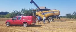 Zdjęcia przedstawia żółty kombajn zbożowy marki New Holland w rozłożoną rurą rozładowczą oraz lekki samochód rozpoznawczo-ratowniczy, które znajdują się na ściernisku. Pomiędzy kombajnem a samochodem strażackim znajdują się dwaj mężczyźni, rozmawiający o bezpieczeństwie podczas żniw. W tle zdjęcia znajdują się lasy i bezchmurne niebo. Zdjęcie wykonane za dnia.