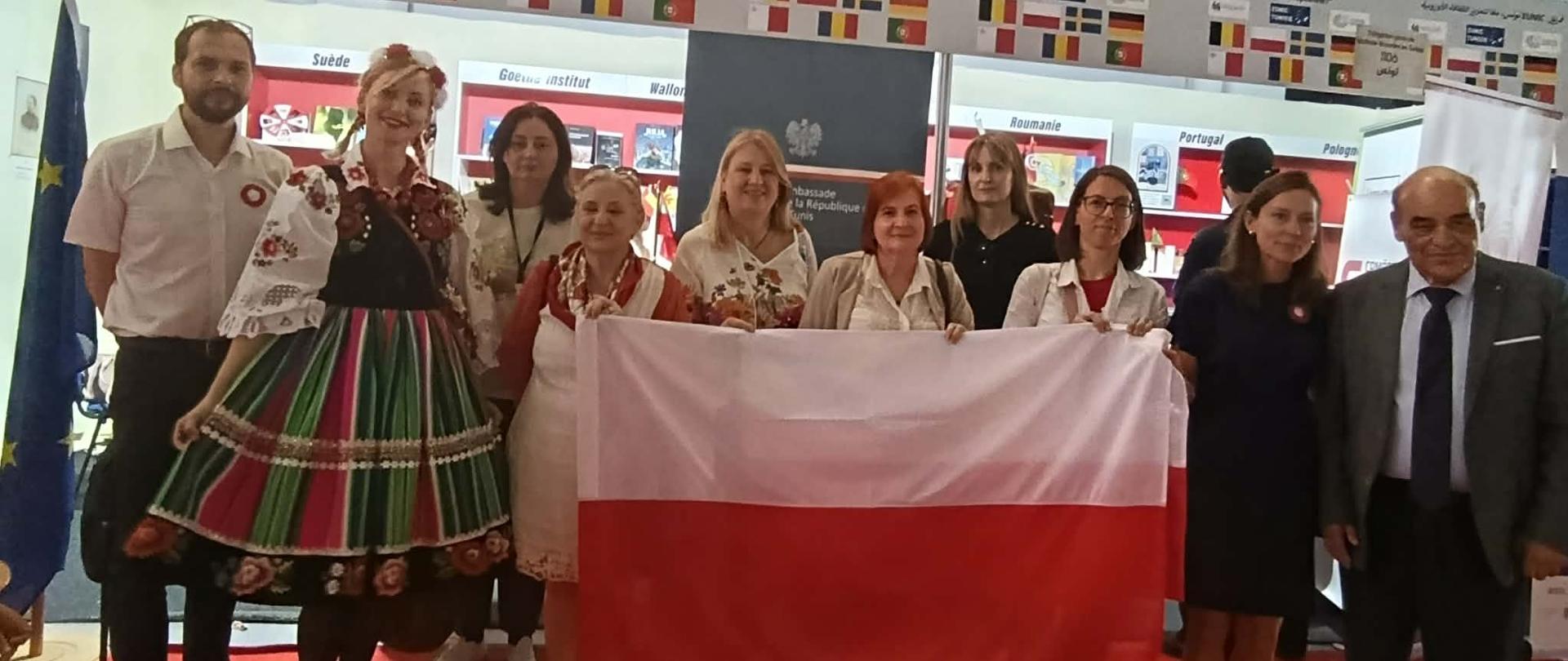 Przedstawiciele Ambasady RP, Konsulatu Honorowego RP, Szkoły Polskiej i Domu Polskiego trzymają w rękach polską flagę, w tle stoisko EUNIC z książkami oraz logo Ambasady RP w Tunisie, po lewej stronie flaga Unii Europejskiej.