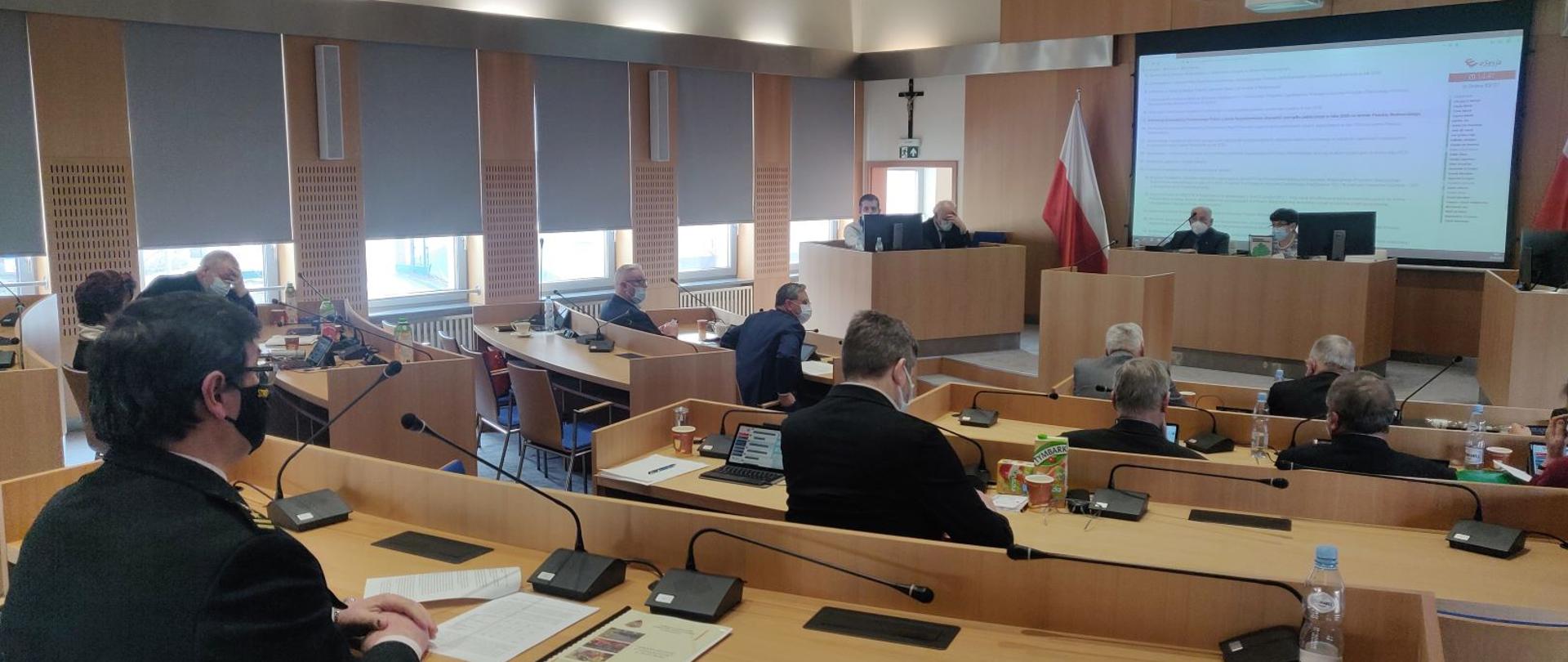 Sala sesyjna. Po lewej stronie Komendant Powiatowy PSP w mundurze wyjściowych siedzący przy stoliku sesyjnym z mikrofonem. W głębi sali widoczni inni uczestnicy. W tle po prawej stronie prezydium Komisji i ekran z prezentacją.
