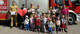 Zdjęcie pokazuje grupę dzieci z opiekunami i strażakami na tle wozu strażackiego.