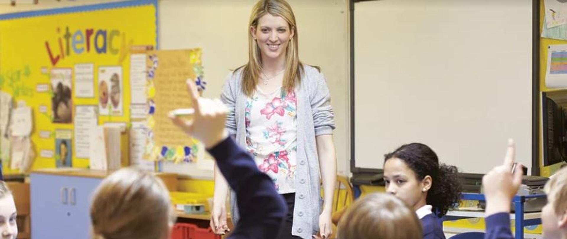 Na zdjęciu grupa uczennic, siedząca tyłem do kadru. Są to dzieci w wieku kilku lat, ubrane w mundurki szkolne. Na przeciw nich stoi nauczycielka. To młoda kobieta z uśmiechem na twarzy. W tle widać tablicę edukacyjną. 