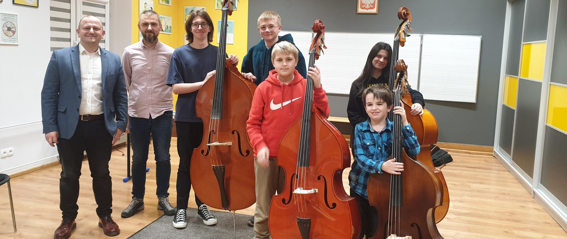 Na zdjęciu znajdują się pedagog prowadzący warsztaty - Wojciech Front oraz nauczyciel kontrabasu i pięcioro uczniów PSM, cztery kontrabasy w sali rytmicznej PSM w kolorach szaro, żółty, biały, na ścianie wisi godło i biała tablica w pięciolinię, po prawej stronie znajduje się szafa w kolorach szaro-żółtym