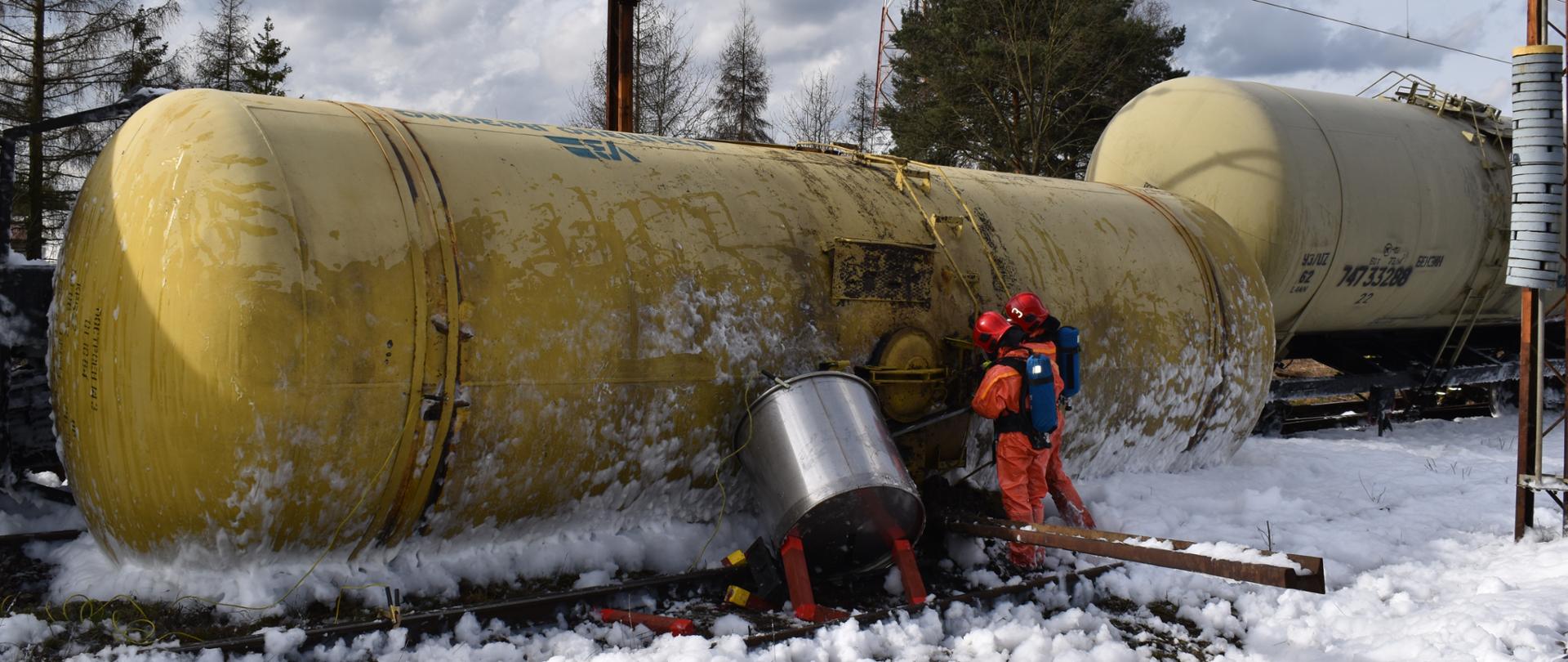 Cysterna leżąca na boku, strażacy w ubraniach specjalistycznych, piana.