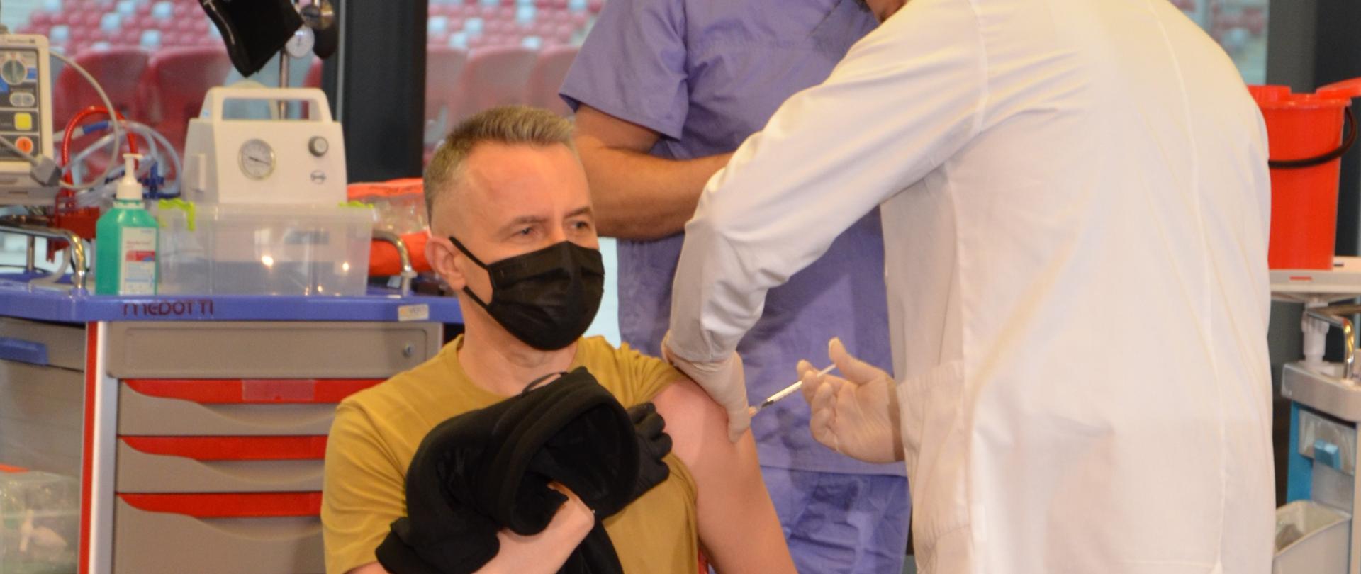 Zdjęcie przedstawia mężczyznę w piaskowej koszulce z podwiniętym lewym rękawem, siedzącego w pomieszczeniu ze sprzętem medycznym. Stojący obok niego mężczyzna w białym kitlu wbija mu w ramie igłę strzykawki. Za nimi stoi jeszcze jeden człowiek w niebieskim kombinezonie. W tle okna i widziane przez nie trybuny.