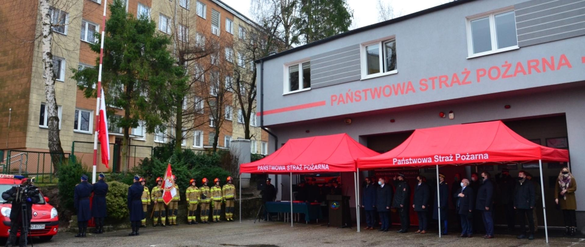 Plac wewnętrzny Komendy Powiatowej w Braniewie. Przy garażach ustawione czerwone namioty a w nich przedstawiciele samorządów i Komendant Wojewódzki. Odbywa się uroczyste wciąganie flagi państwowej na maszt. W tle budynek mieszkalny.