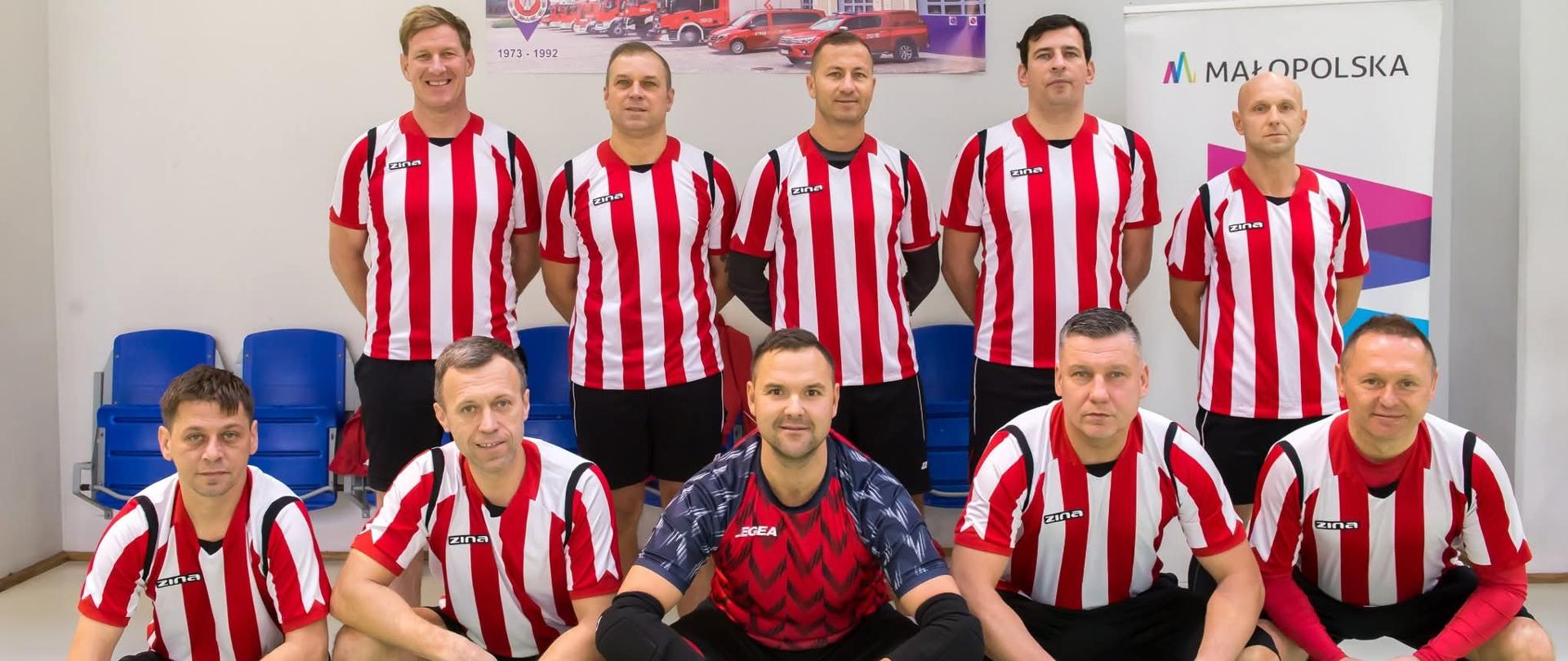 Grupowe zdjęcie drużyny sportowej składającej się z jedenastu mężczyzn. Dziesięciu z nich ma na sobie biało-czerwone, pionowo paskowane koszulki i czarne spodenki, a jeden – w środku pierwszego rzędu – nosi czarno-czerwoną koszulkę bramkarską oraz ochraniacze na kolanach. Pięciu zawodników stoi z tyłu, a sześciu kuca z przodu. Zdjęcie wykonano w hali sportowej; w tle widoczne są niebieskie krzesełka oraz baner z napisem „65 lat Krakowskiej Szkoły Pożarniczej” i fotografia budynku z wozami strażackimi. Po prawej stronie stoi roll-up z logo „Małopolska