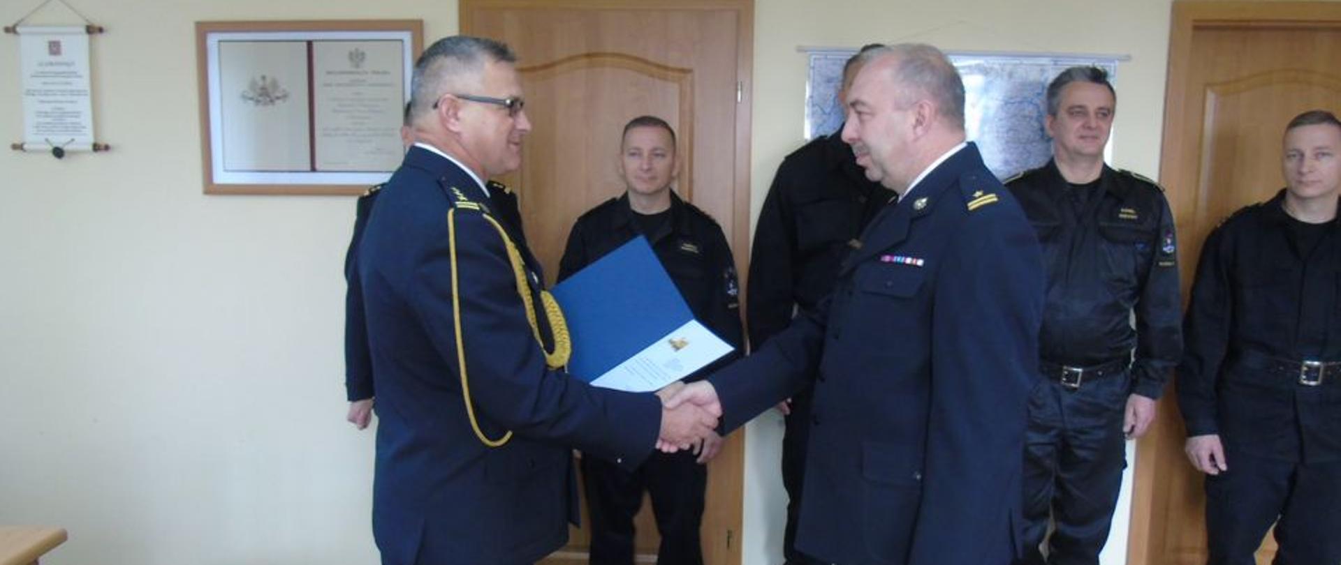 Na zdjęciu na pierwszym planie stoją strażacy w umundurowaniu wyjściowym ( gabardyna)
stoją naprzeciw siebie, podają sobie dłonie, jeden ze strażaków przekazuje list gratulacyjny, na drugim planie stoją w szeregu strażacy ubrani w umundurowanie dowódczo-sztabowe
