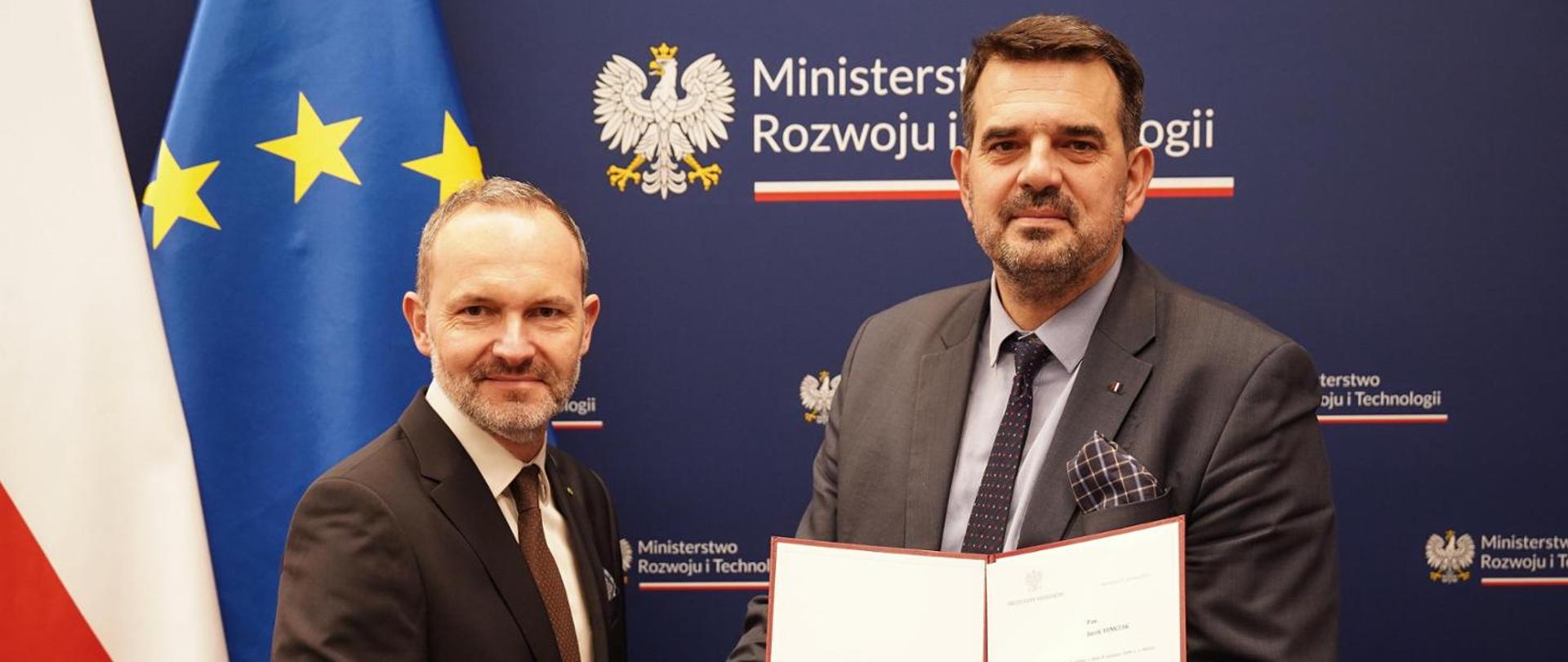 Na zdjęciu widać ministra Krzysztofa Hetmana, który wręcza akt powołania Jackowi Tomczakowi na wiceministra rozwoju. Z tyłu za ministrami baner MRiT, po lewej stronie flaga Polski i Unii