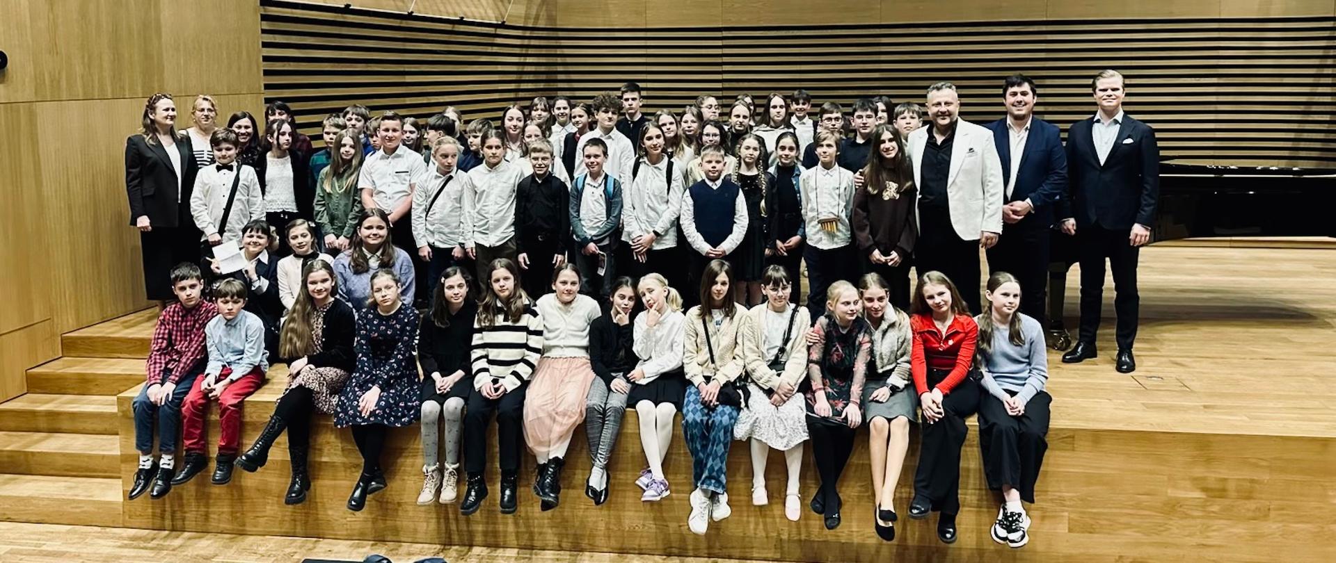 Kolorowe zdjęcie na którym znajduje się grupa ok 60 osób, młodzieży rozmieszczonej w 4 rzędach. Pierwszy rząd osób siedzi na brzegu sceny a kolejne usytuowane są za nimi, którym towarzyszą nauczyciele i soliści koncertu. Zdjęcie zrobione na sali koncertowej PSM Jastrzębie. 