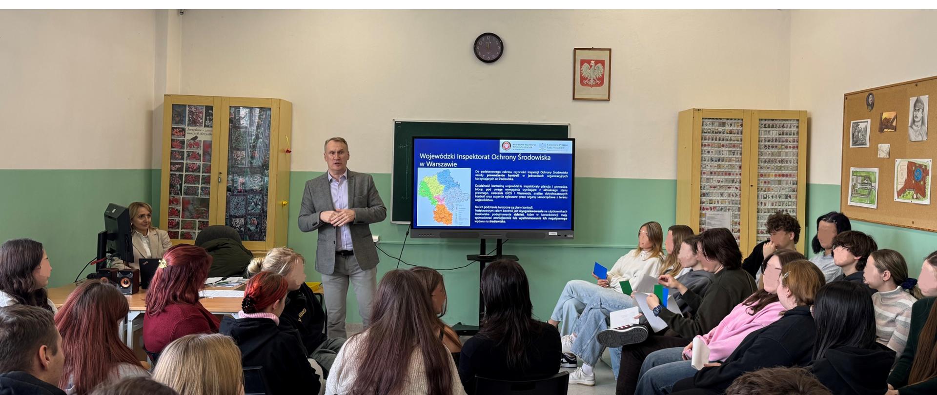 Inspektor Wojewódzkiego Inspektoratu Ochrony Środowiska przedstawia w szkolnej klasie prezentację dotyczącą Wojewódzkiego Inspektoratu Ochrony Środowiska. Przed nim siedzi grupa uczniów trzymając kolorowe kartki. W tle widać szkolne gabloty, tablicę i godło Polski na ścianie.