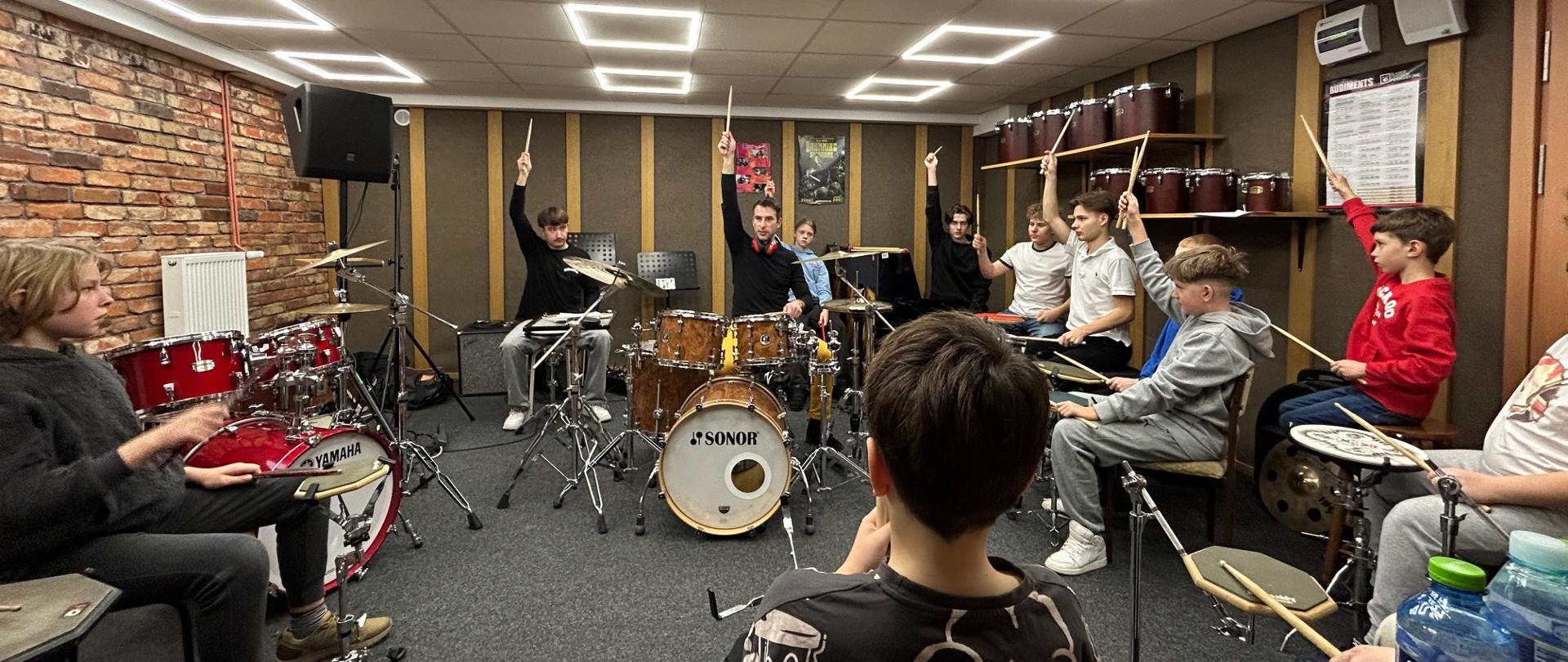 Grupa uczniów siedzących przy zestawach perkusyjnych w sali muzycznej, ćwiczących pod kierunkiem prowadzącego warsztaty.