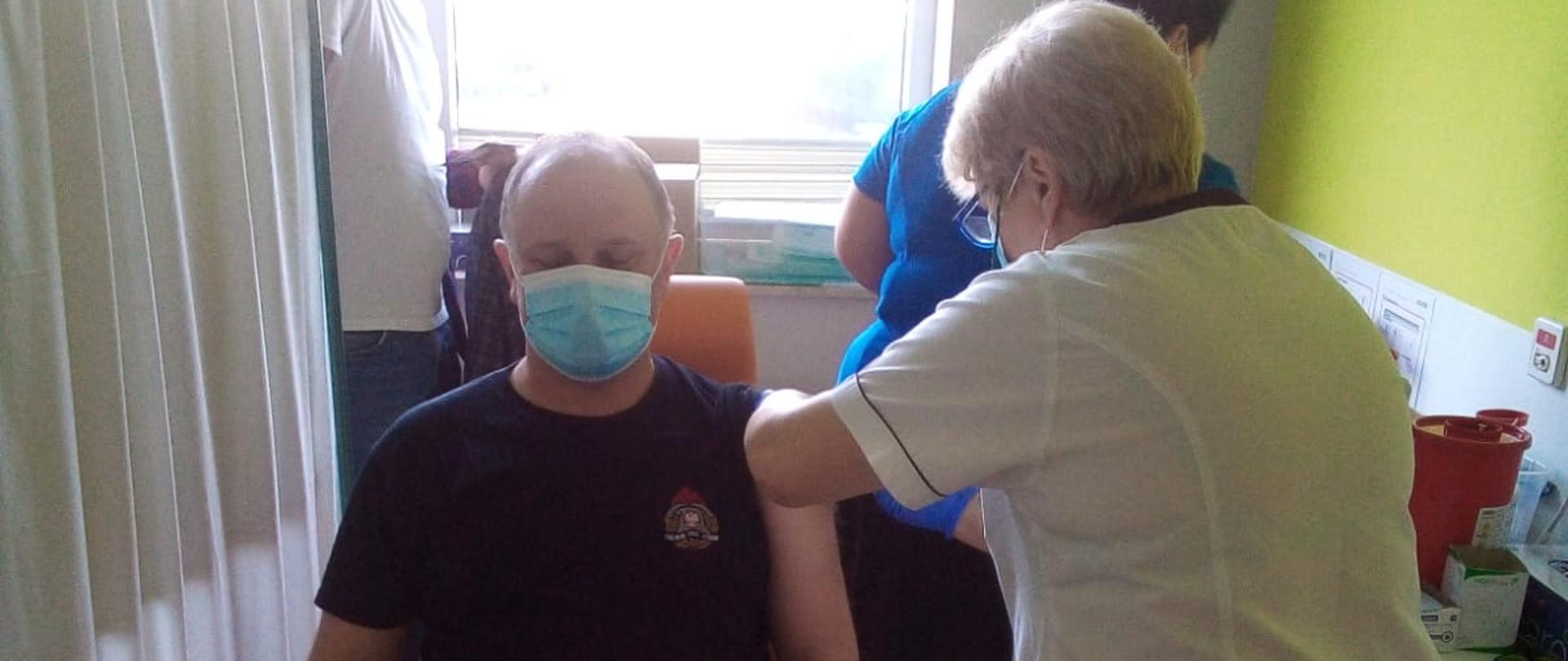Mężczyzna ubraną w czarną koszulkę strażacką z krótkim rękawem, na twarzy ma ubrana maseczkę chirurgiczną koloru biało-niebieską, siedzi on na krześle koloru pomarańczowego w gabinecie szpitalnym, po prawej stronie ma parawan w kolorze białym. Na lewej ręce ma podwinięty rękaw do góry. Obok stoi pielęgniarka ubrana w biały fartuch oraz czarne spodnie i podaje szczepionkę w lewe ramie strażaka. Szczykawka jest wbita w ramię. W tle stoi mężczyzna ubrany w białą koszulkę w krótkim rękawem, na twarzy ma ubrana maseczkę chirurgiczną koloru biało-niebieską. Obok stoi druga pielęgniarka ubrana w niebieską koszulę