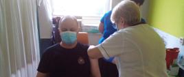 Mężczyzna ubraną w czarną koszulkę strażacką z krótkim rękawem, na twarzy ma ubrana maseczkę chirurgiczną koloru biało-niebieską, siedzi on na krześle koloru pomarańczowego w gabinecie szpitalnym, po prawej stronie ma parawan w kolorze białym. Na lewej ręce ma podwinięty rękaw do góry. Obok stoi pielęgniarka ubrana w biały fartuch oraz czarne spodnie i podaje szczepionkę w lewe ramie strażaka. Szczykawka jest wbita w ramię. W tle stoi mężczyzna ubrany w białą koszulkę w krótkim rękawem, na twarzy ma ubrana maseczkę chirurgiczną koloru biało-niebieską. Obok stoi druga pielęgniarka ubrana w niebieską koszulę.