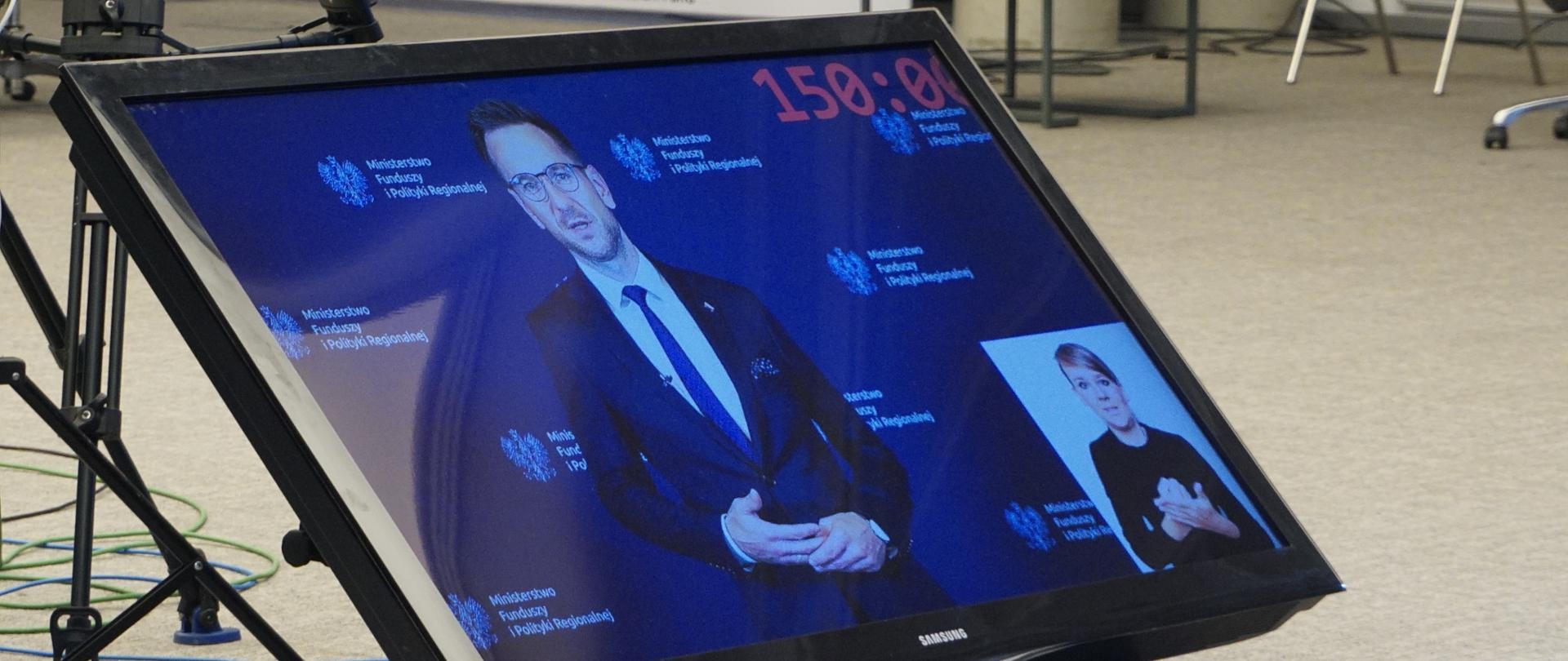 Na monitorze, który stoi na podłodze, zdjęcie wiceministra Waldemara Budy