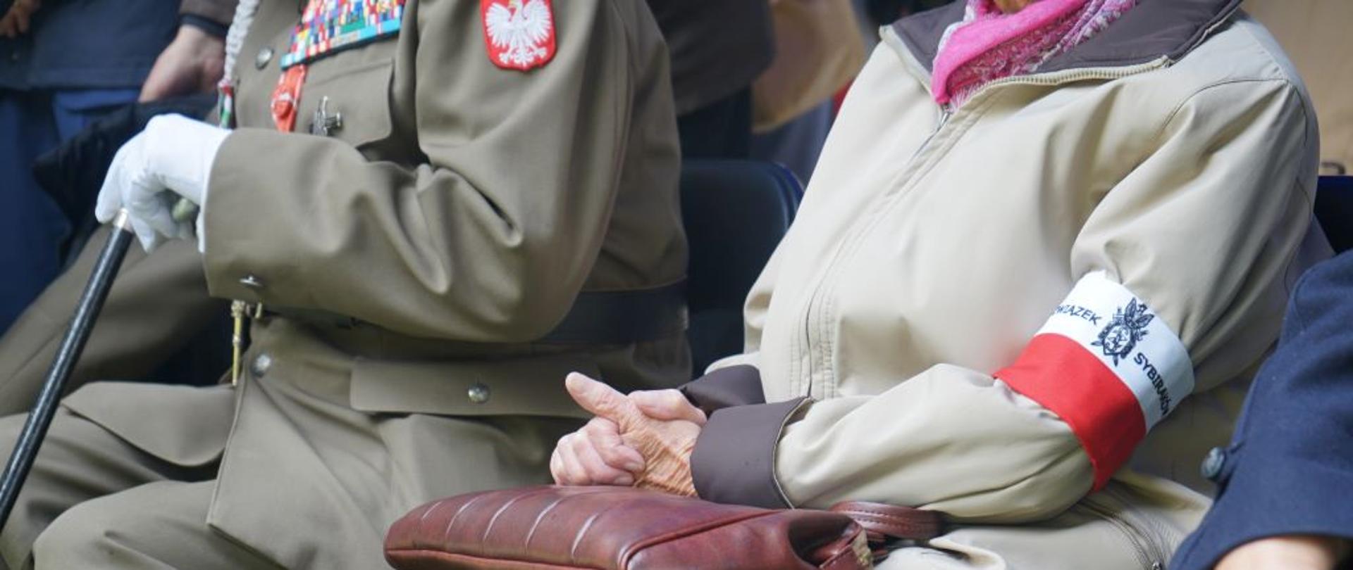 osoby siedzące na krzesłach, nie widać twarzy, widoczna opaska biało czerwona założona na rękaw kurtki, oraz zielony mundur z medalami
