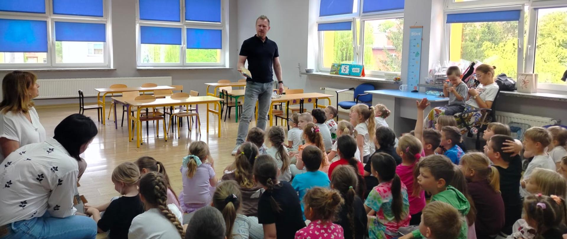W przestronnej sali z dużymi oknami i niebieskimi roletami grupa dzieci siedzi na podłodze i słucha Inspektora stojącego pośrodku pomieszczenia. Inspektor trzyma w ręce worek z nasionami. Po bokach znajdują się opiekunowie oraz stoliki i krzesła szkolne. Atmosfera przypomina spotkanie edukacyjne lub warsztaty.