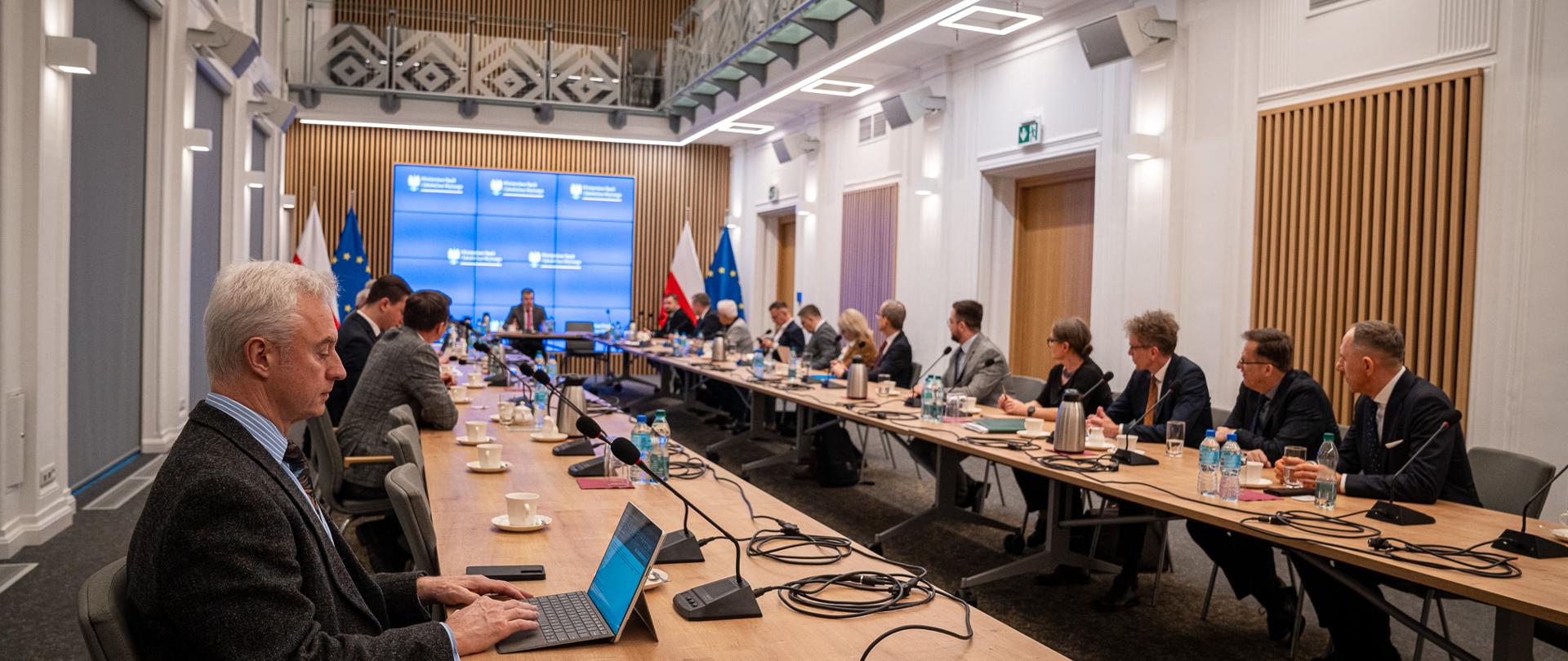 Widok od tyłu na salę, przy ustawionych w czworokąt stołach siedzi wiele osób.