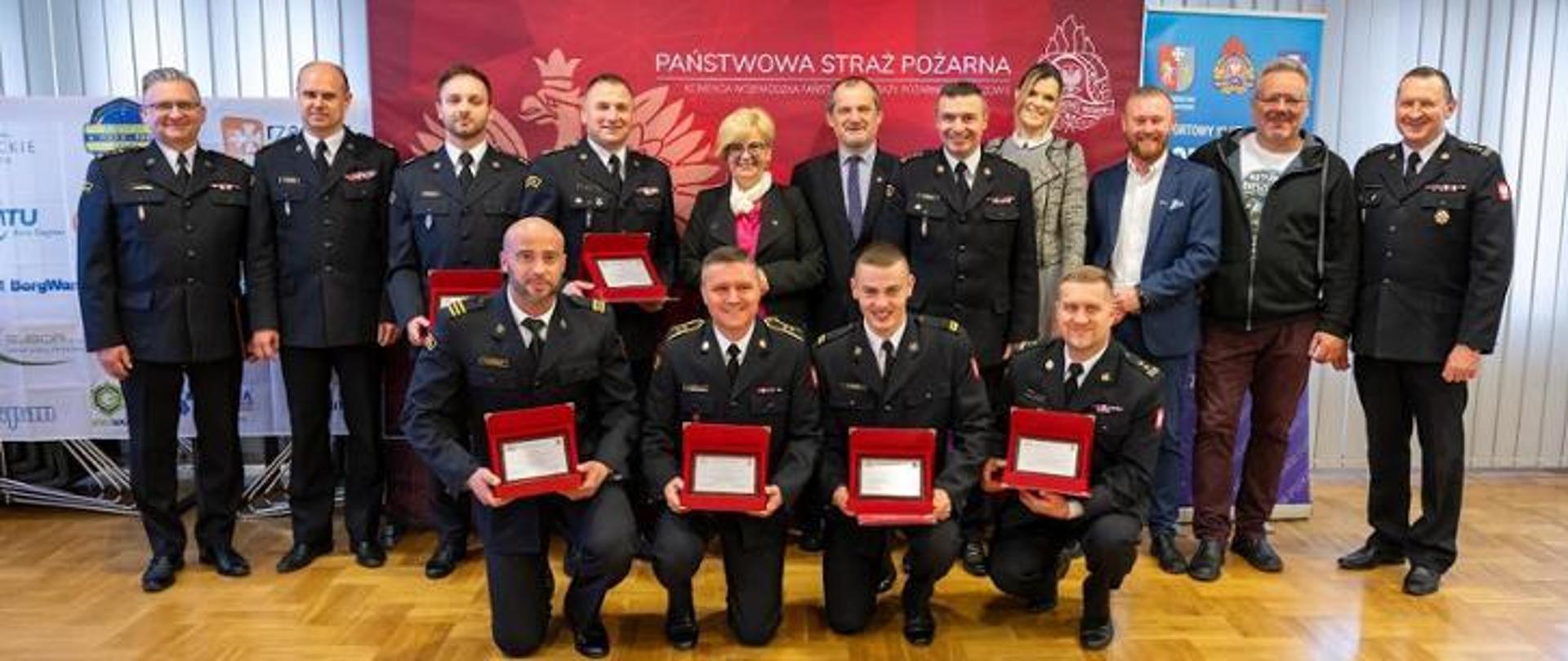 Fotografia wykonana podczas spotkania ze strażakami sportowcami w komendzie wojewódzkiej Państwowej Straży Pożarnej w Rzeszowie. Na zdjęciu reprezentacja strażaków z województwa podkarpackiego z wyróżnieniami za osiągane wyniki na szczeblu krajowym i międzynarodowym. W pierwszym rzędzie stoją również zaproszeni goście na uroczystości. Funkcjonariusze ubrani są w umundurowanie wyjściowe Państwowej Straży Pożarnej koloru granatowego. Strażacy sportowcy trzymają w rękach wyróżnienia w postaci pamiątkowych tablic. Zdjęcie wykonane na tle bajeru z napisem Państwowa Straż Pożarna.