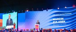 Na podwyższeniu przed wielkim ekranem z napisem na niebieskim tle KNM 2026 Opening Ceremony - Marcin Kulasek stoi minister Kulasek i mówi do mikrofonu na stojaku, z boku stoi kobieta w czerwonej sukni.