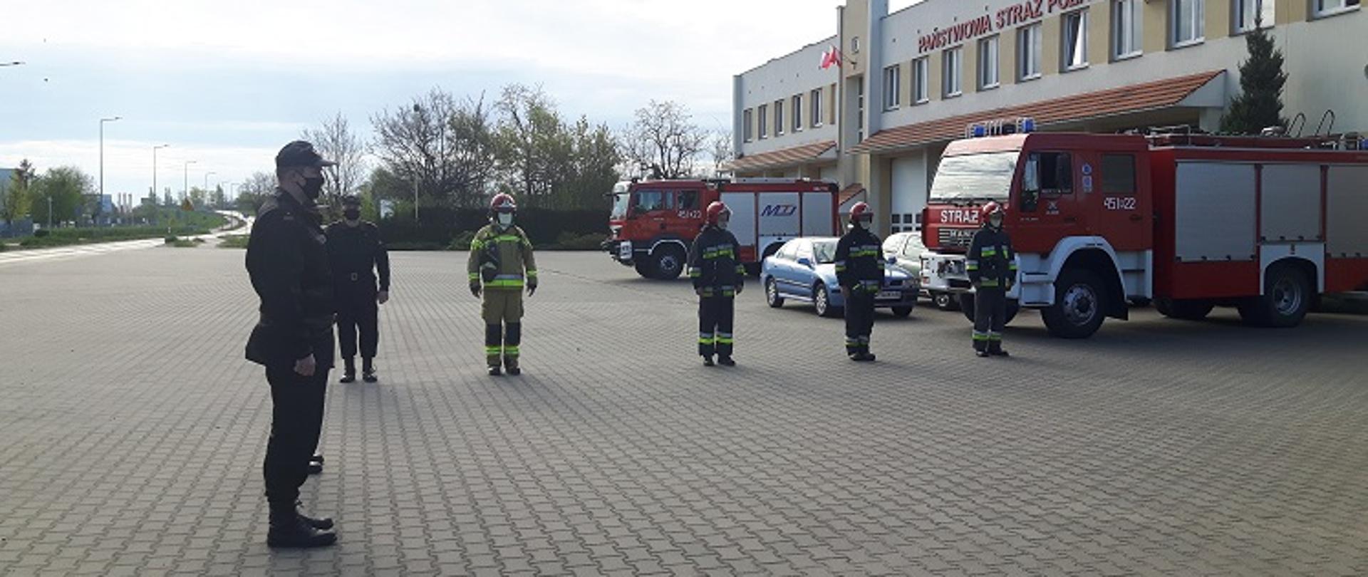 Na tle budynku Komendy ustawione samochody strażackie. Funkcjonariusze stoją na baczność podczas uroczystego apelu.